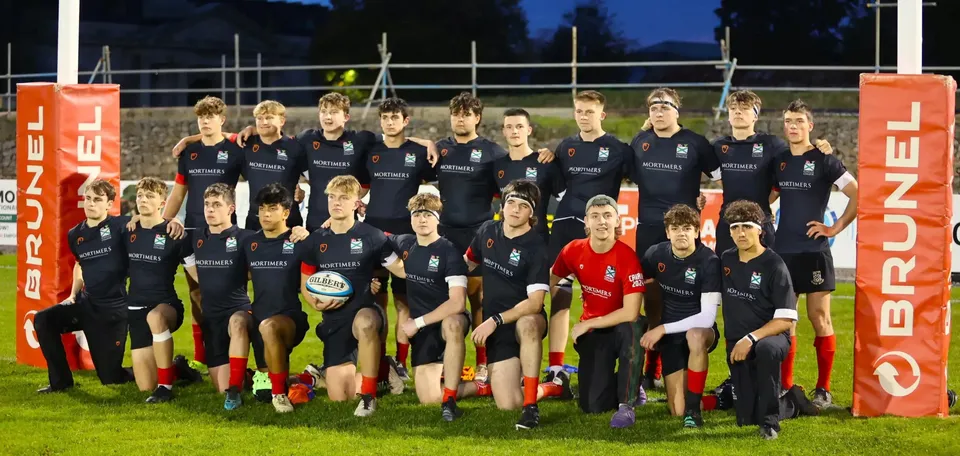 Un gruppo di giovani atleti in divisa si trova sul campo sportivo del Plymouth College di notte sotto i riflettori.