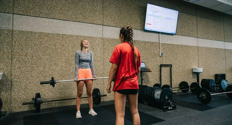 Due donne in abbigliamento sportivo sono in piedi nella palestra della Mission Secondary School davanti ad attrezzi da allenamento e a uno schermo.