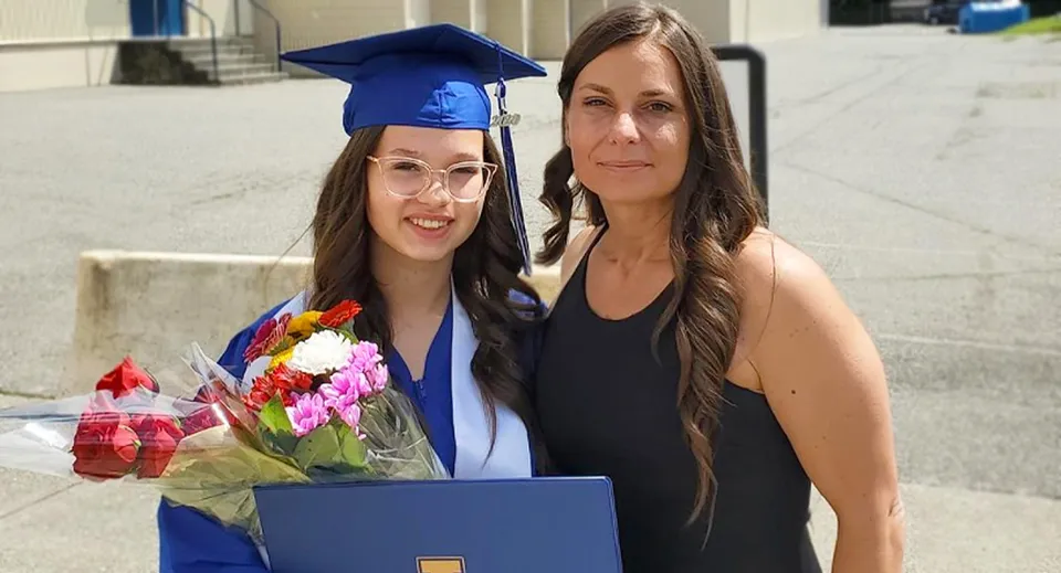 Una studentessa della Aldergrove Community Secondary School in toga blu da diploma sta in piedi accanto a una donna più anziana con dei fiori davanti all'edificio scolastico.