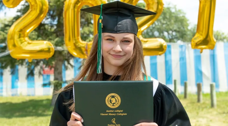 Una giovane donna in toga e tocco di laurea tiene un diploma davanti a numeri fatti di palloncini gialli nel campus dell'Institut Collégial Vincent Massey Collegiate.