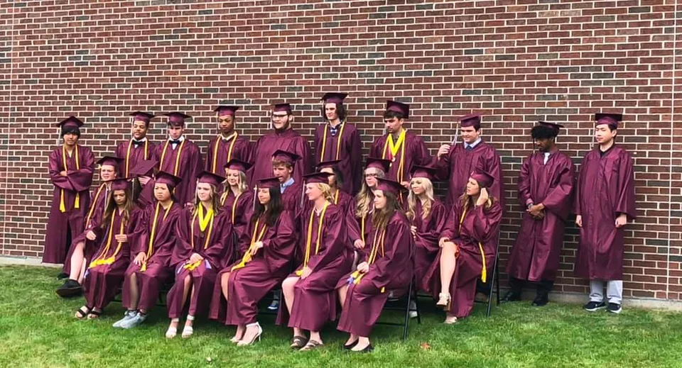 Diplomati e diplomate della N.E.W. Lutheran High School in piedi in toga davanti a una parete in mattoni mentre festeggiano il diploma.