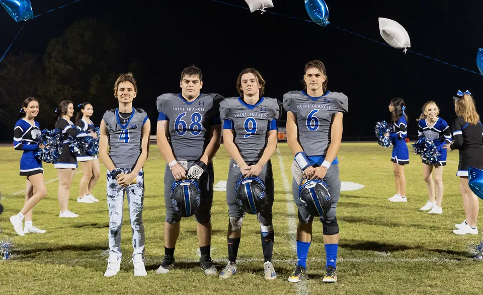 Un gruppo di cheerleader della Saint Francis Catholic Academy si trova di sera sul campo da football sotto luci brillanti.