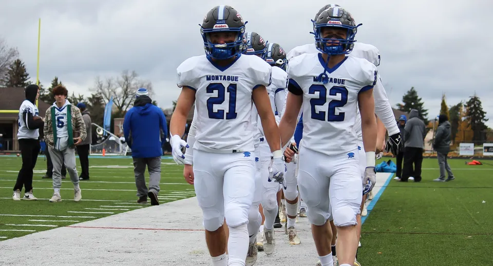Due giocatori di football della Montague Regional High School in maglia bianca corrono insieme sul campo da gioco.