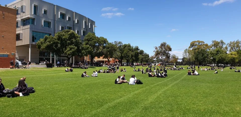 Molti studenti sono seduti su un prato verde davanti all'edificio a più piani della Adelaide High School.