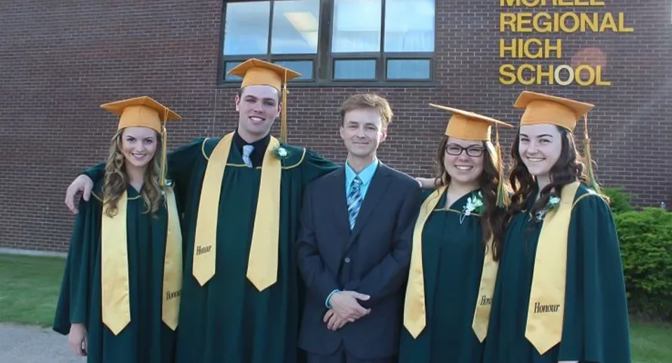 Un gruppo di diplomati e diplomate della Morell Regional High School in toga verde sta davanti all'edificio scolastico insieme a un amministratore della scuola.