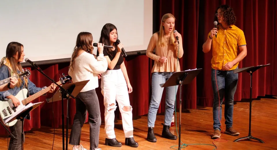 Un gruppo di studenti della Central Christian School prova sul palco davanti a un sipario rosso con i microfoni.