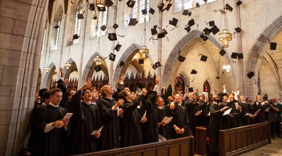 I diplomati della Cranbrook Schools in toga nera lanciano i loro tocchi in aria all'interno di un grande salone in pietra.