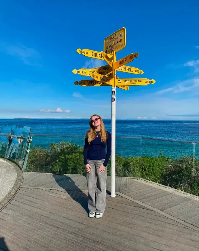 Studentessa sotto un cartello segnaletico giallo con destinazioni del mondo davanti al mare in Nuova Zelanda