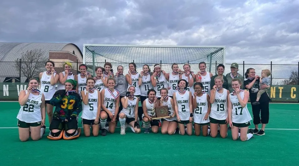 Un gruppo di giovani sportive posa sul campo di hockey del Montpelier Roxbury Public School District sotto un cielo nuvoloso.