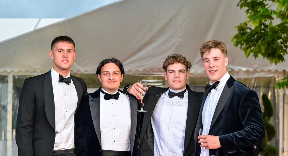Quattro studenti della Sherborne School in abito formale posano insieme davanti a una tenda e a un ambiente verde.