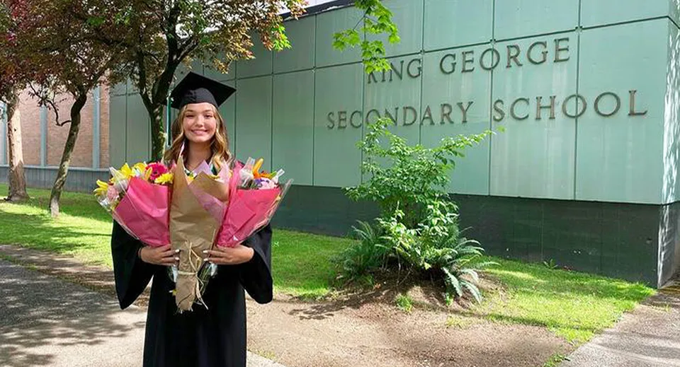 Una diplomata è in piedi davanti all'edificio verde della King George Secondary School e tiene un mazzo di fiori.