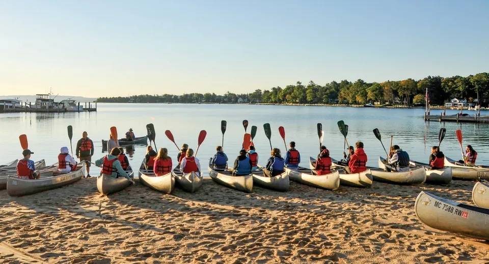Studenti del Coláiste Éamann Rís pagaiano in canoa con abiti tradizionali su un lago calmo circondato dal bosco.