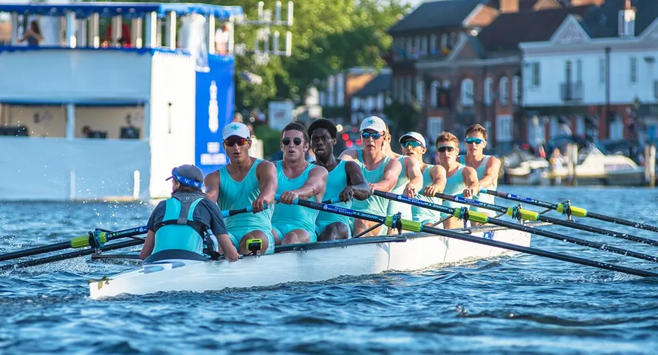 Un gruppo di canottieri con divise turchesi coordinate siede su una barca da canottaggio in acqua davanti a uno scenario suggestivo nel campus dell'Eton College.