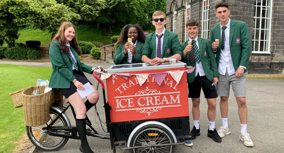 Studenti della Woodhouse Grove School sono radunati intorno a un carretto del gelato su un prato davanti a un edificio in mattoni.