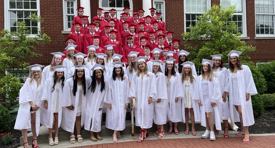 Un grande gruppo di studenti della Beechwood Sacred Heart School indossa toghe rosse e bianche davanti a un edificio in mattoni.