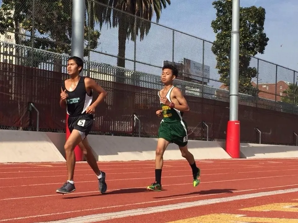 Due atleti della Downtown Magnet High School corrono sulla pista da atletica rossa con una recinzione ed edifici sullo sfondo.