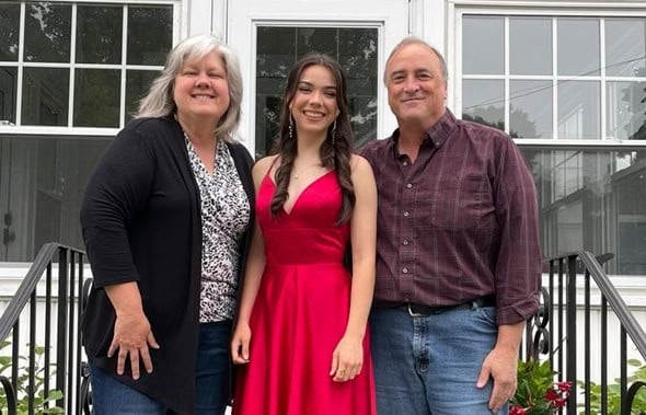 Joven con vestido rojo posa sonriente junto a dos adultos ante una casa