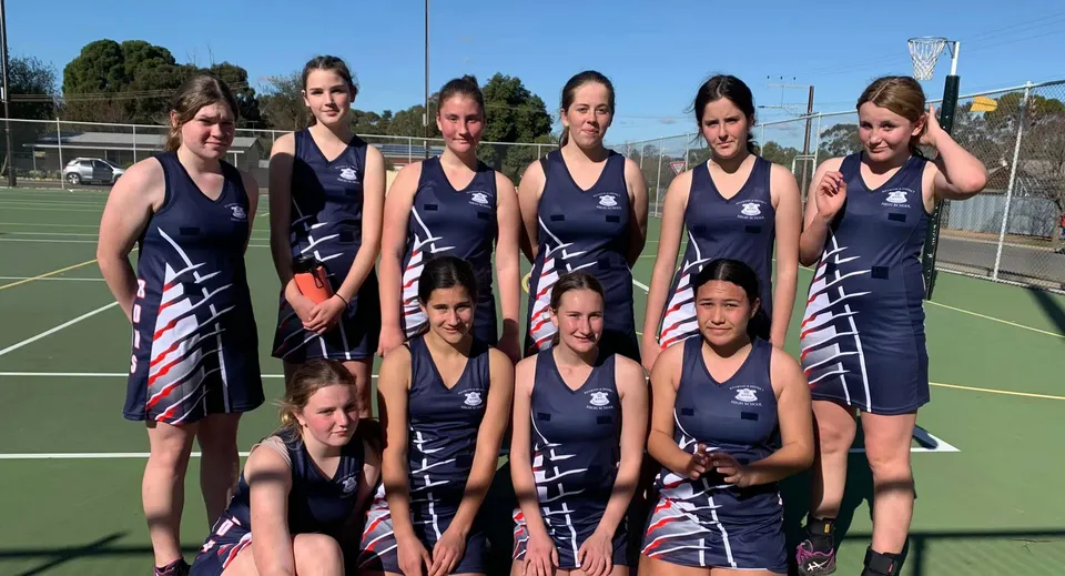 Un gruppo di giovani atlete posa sul campo sportivo della Riverton and District High School davanti ad alberi ed edifici.