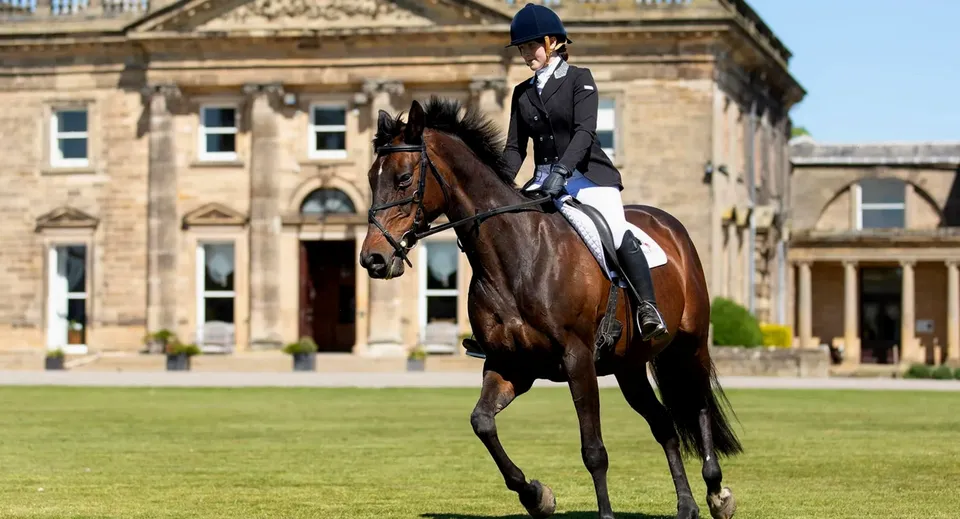 Una persona in tenuta da equitazione formale cavalca un cavallo baio su un campo erboso davanti a un edificio storico nel campus della Queen Mary's School.