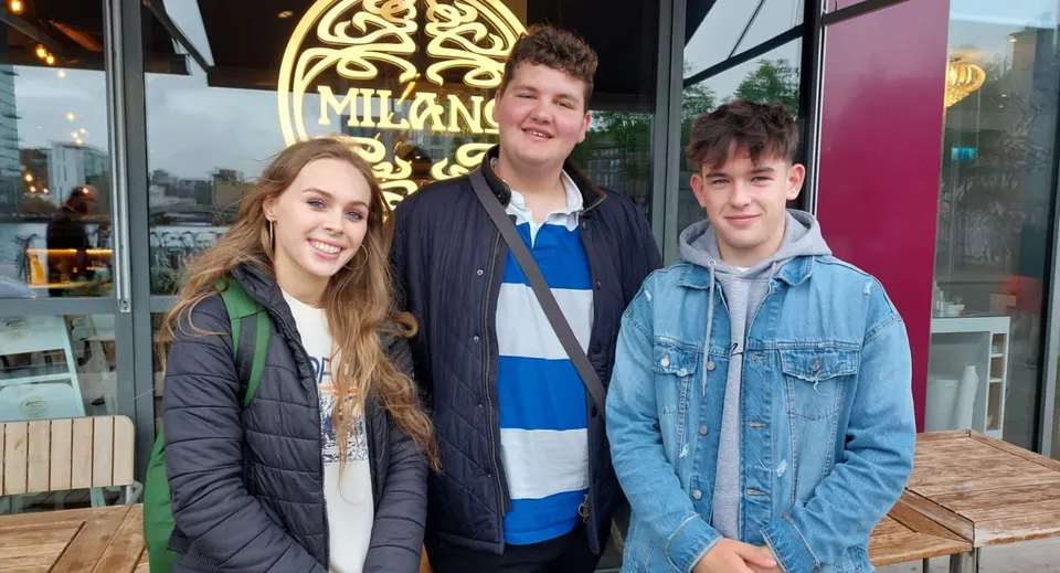 Tre studenti della Newtown School sono in piedi davanti a un bar con un cartello decorativo sullo sfondo.