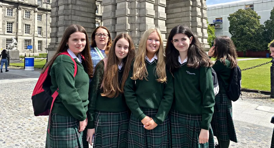 Quattro studentesse della St. Louis Community School in divisa scolastica verde posano all'aperto davanti a un edificio storico in pietra.