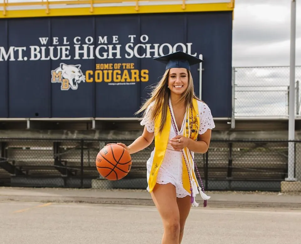 Una diplomata della Mount Blue High School è in toga e con un pallone da basket davanti a un simbolo della scuola.