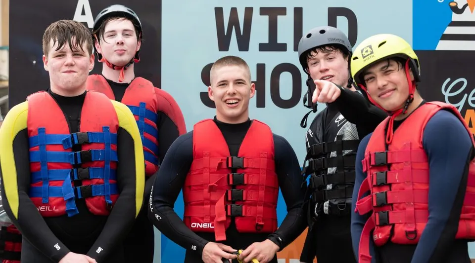 Un gruppo di studenti del Stewart’s Melville College indossa giubbotti salvagente e caschi davanti a un cartello con la scritta WILD 100.