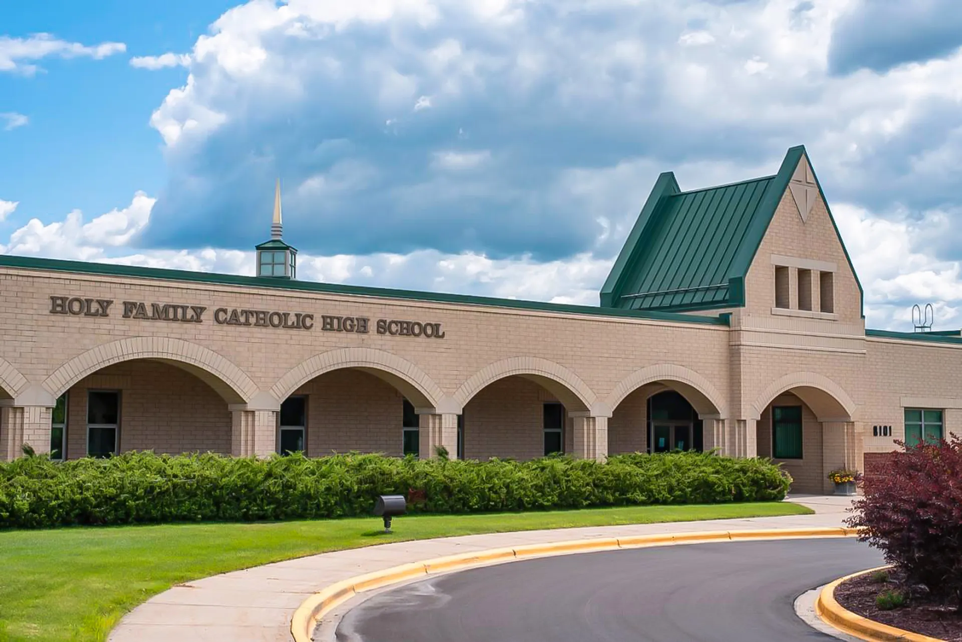 Il grande edificio in mattoni della Holy Family Catholic High School con portale ad arco e tetto verde a torretta è circondato dal verde e da un cielo nuvoloso.