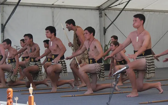 Un grupo de hombres con vestimenta tradicional realiza una danza sincrónica sobre el escenario en el campus de la Whangamata Area School.