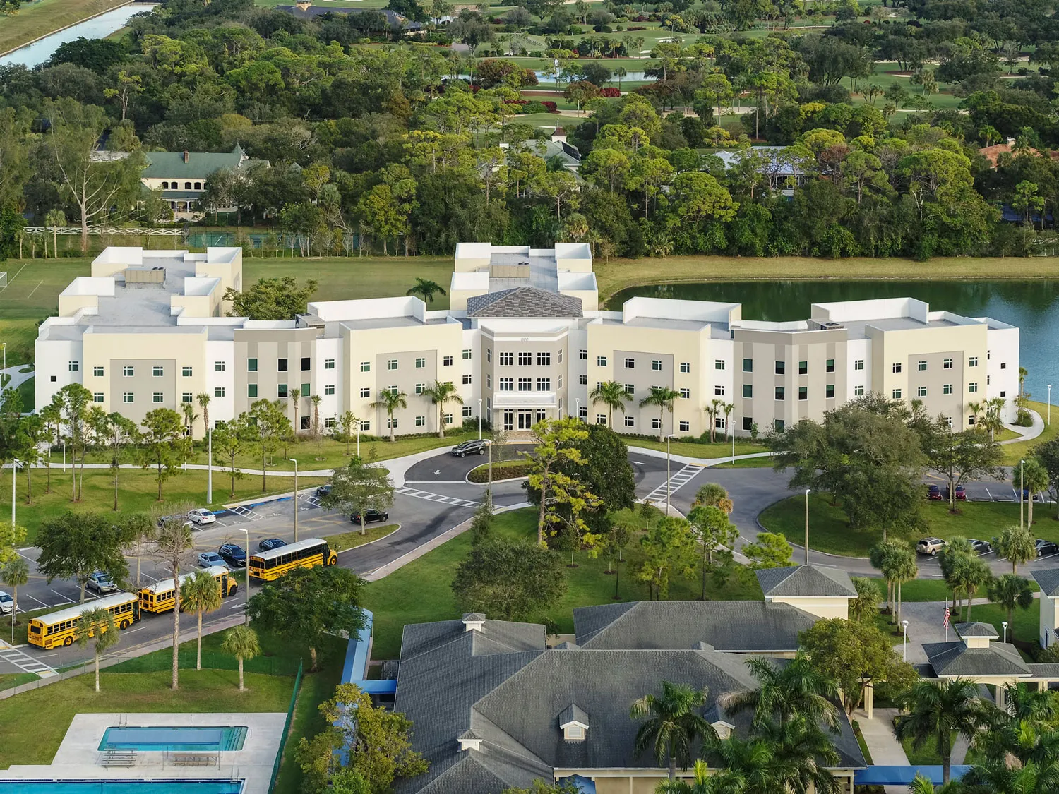 Vista aérea del edificio principal de North Broward Preparatory School rodeado de palmeras, un lago y autobuses escolares.