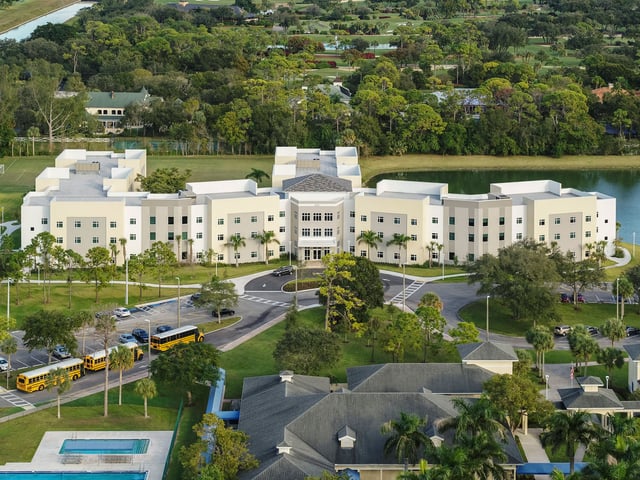Luftaufnahme des großen Schulgebäudes der North Broward Preparatory School mit gelben Schulbussen, Palmen und einem See.