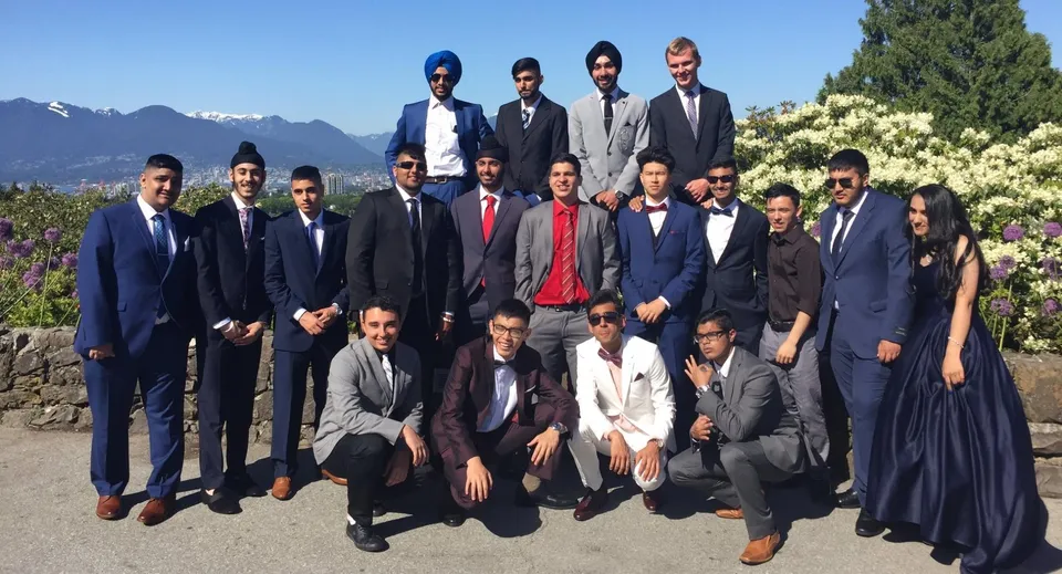 Un gruppo di uomini in abito formale posa all'aperto con un paesaggio montano sullo sfondo nel terreno della Enver Creek Secondary School.