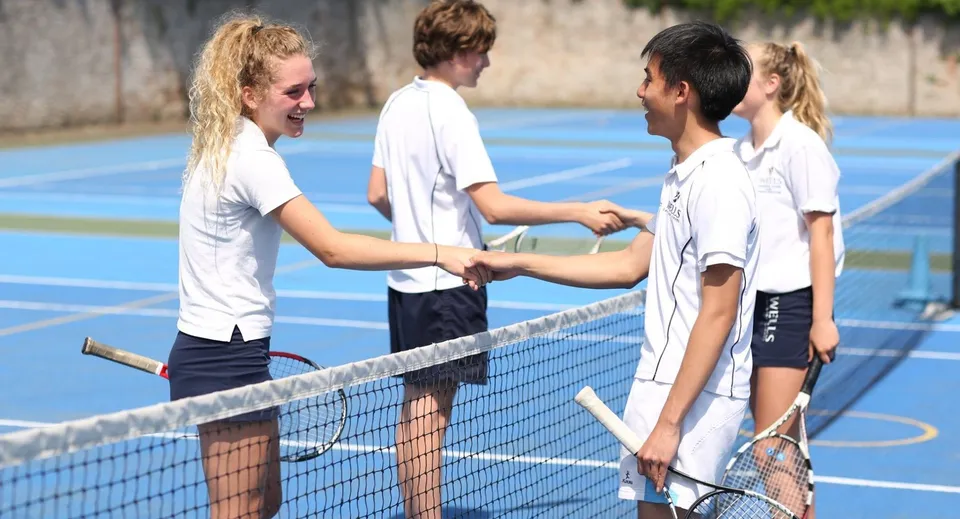 Studenti della Wells Cathedral School si stringono la mano su un colorato campo da tennis all'aperto.