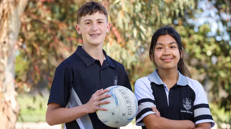 Due studenti della Adelaide High School sono all'aperto e tengono un pallone da rugby davanti agli alberi.