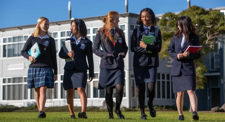 Cinque studentesse in divisa sono insieme nel campus della New Plymouth Girls’ High School con un edificio sullo sfondo.
