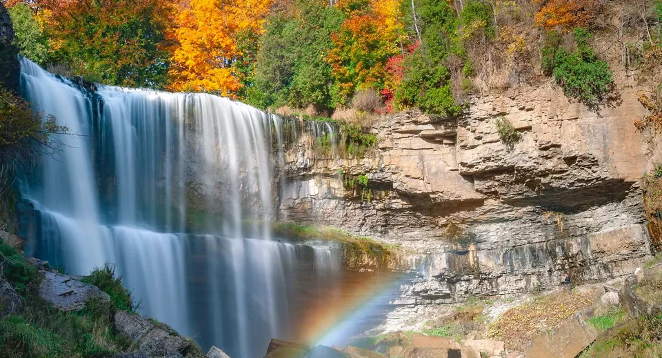 Una cascata scorre su una parete rocciosa con fogliame autunnale sullo sfondo nel campus della Innisdale Secondary School.