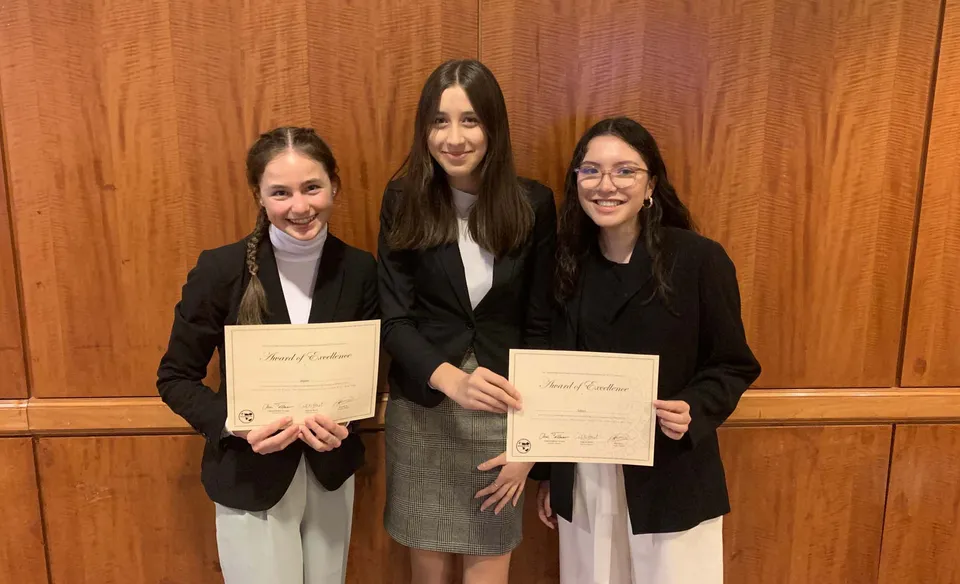 Tre donne in abbigliamento formale tengono in mano dei certificati davanti a una parete rivestita in legno nel campus della Bellarmine Preparatory School.