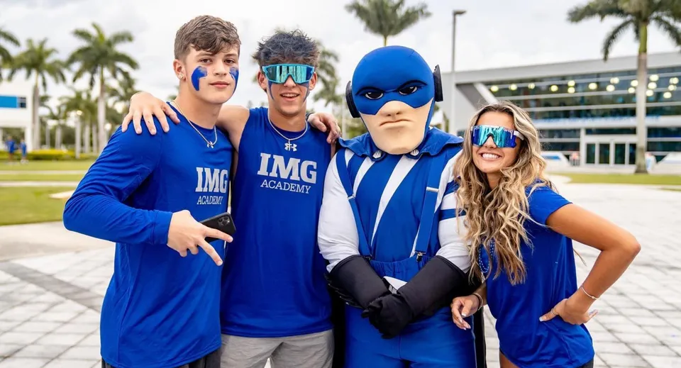Un gruppo di quattro studenti indossa divise blu della IMG Academy davanti a un edificio con palme.