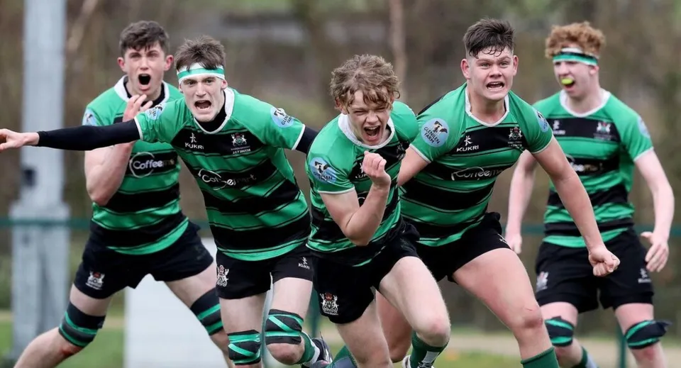 Studenti della Bandon Grammar School giocano a rugby insieme su un campo erboso con alberi sullo sfondo.