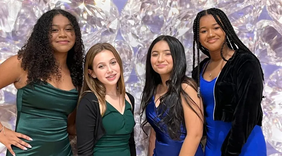 Quattro studentesse della Our Savior New American School posano sorridenti con abiti eleganti davanti a uno sfondo decorato.