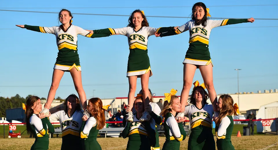 Studenti e studentesse della Kenston Forest School eseguono uno stunt di cheerleading sul campo sportivo sotto un cielo limpido.
