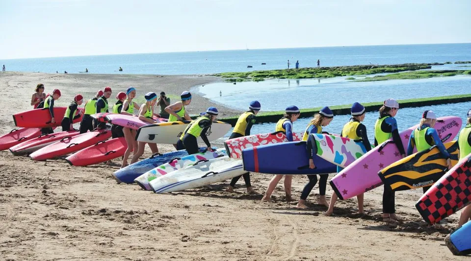Un gruppo di studenti della St. John’s School sulla spiaggia indossa giubbotti di salvataggio colorati ed è in piedi davanti al mare.