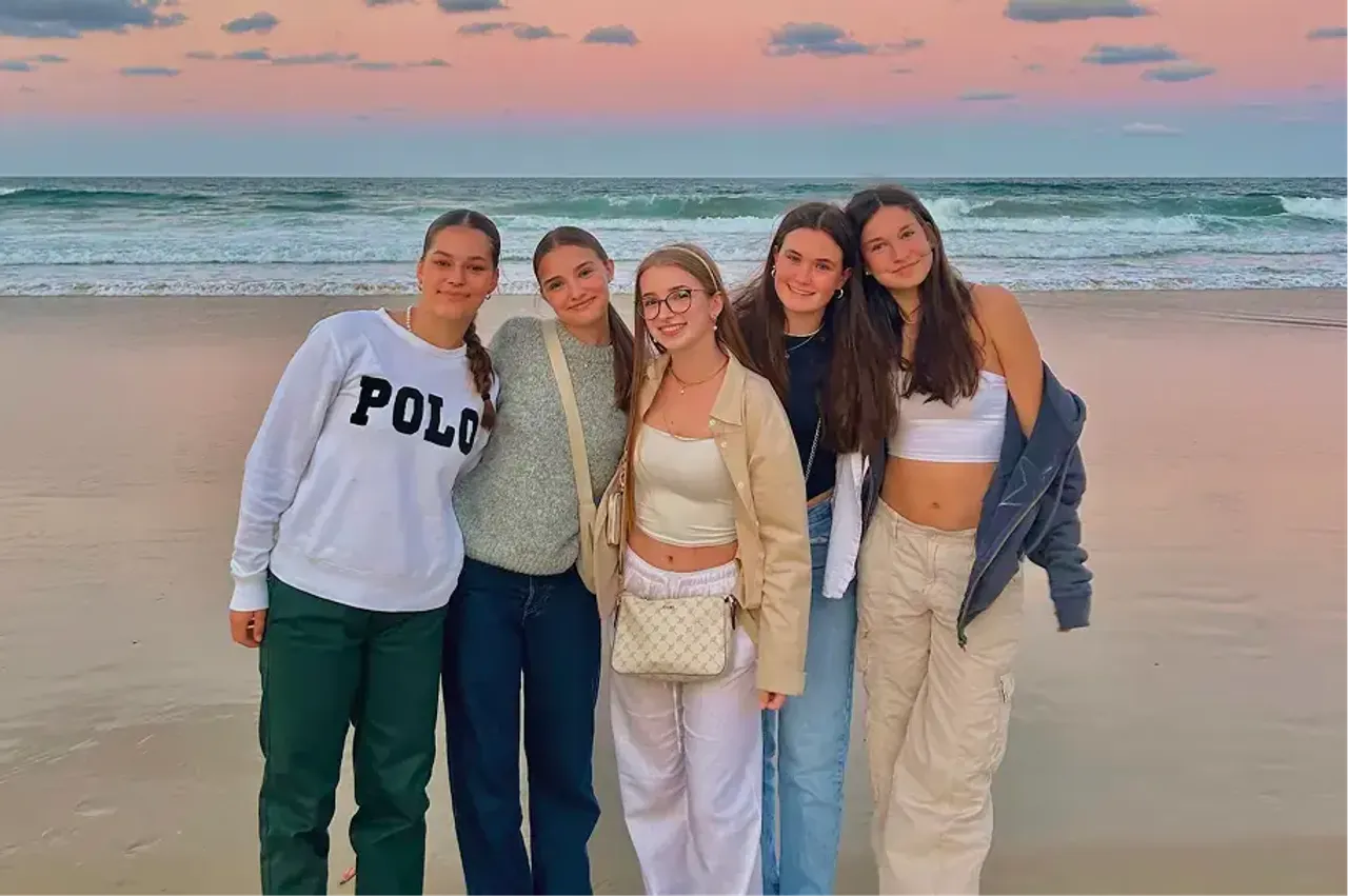 Cinque amiche posano sorridenti su una spiaggia al tramonto con il cielo rosa