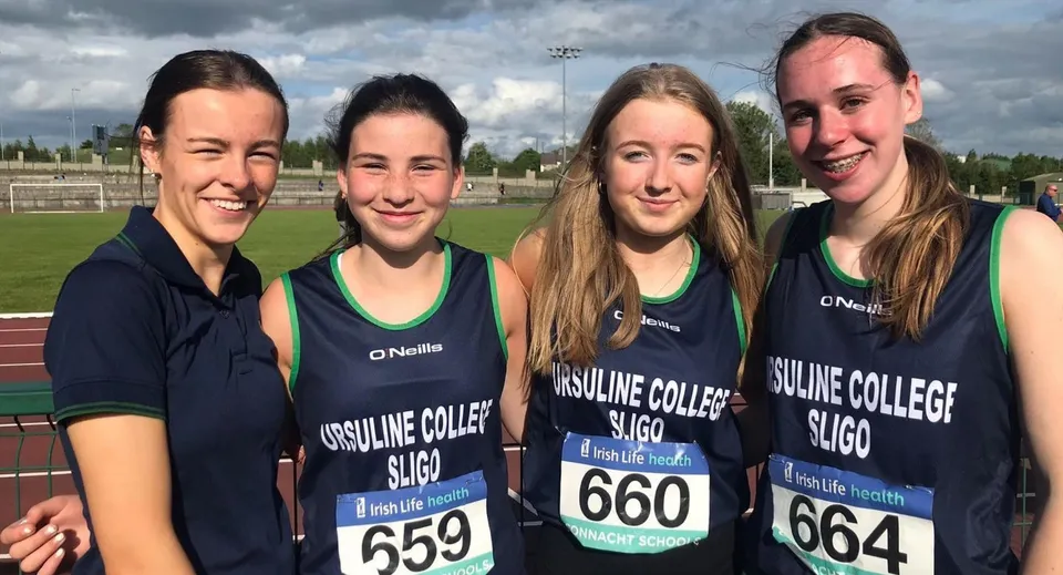 Quattro studentesse dell'Ursuline College Sligo su un campo sportivo sotto un cielo nuvoloso, indossando divise sportive abbinate.