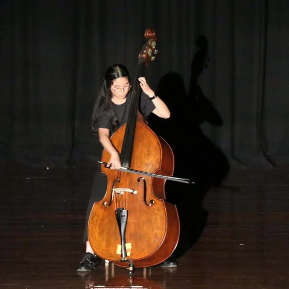 Una studentessa della Riverside Girls High School suona il contrabbasso su un palco davanti a un sipario scuro.
