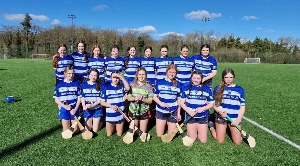 Un gruppo di studentesse del Comeragh College posa insieme in maglie a strisce blu e bianche su un campo da calcio davanti a un bosco e al cielo blu.