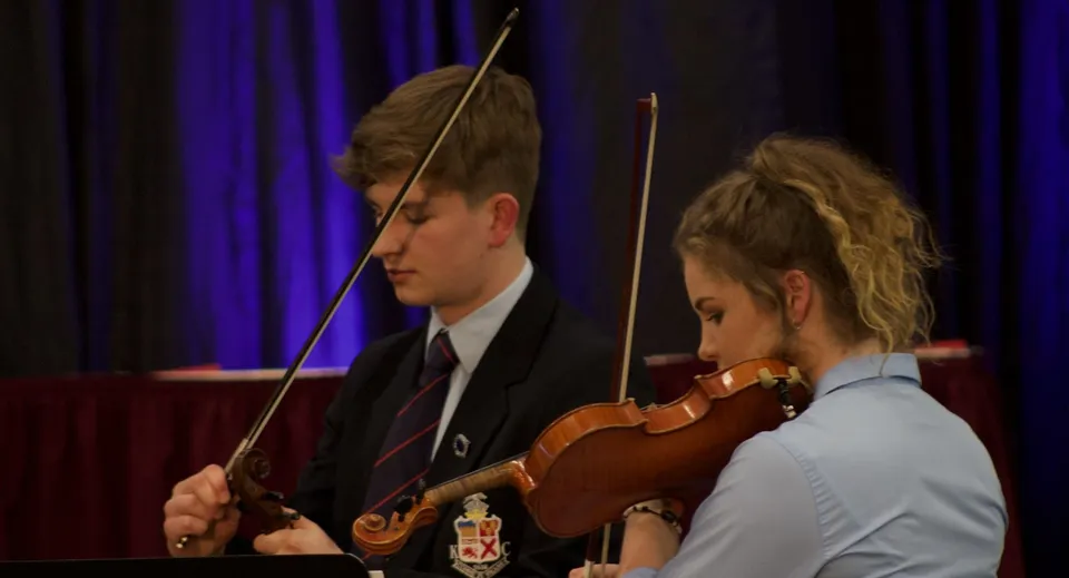 Due studenti del Kilkenny College si esibiscono insieme con violino e archetto su un palco con il sipario rosso.