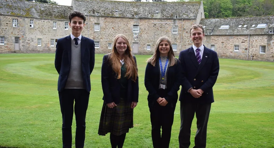 Quattro studenti in uniforme scolastica sono in piedi su un prato davanti a uno storico edificio in pietra di Gordonstoun.