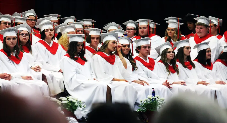 I diplomati della St. Thomas Aquinas High School siedono insieme su un palco indossando toghe bianche con sciarpe rosse.
