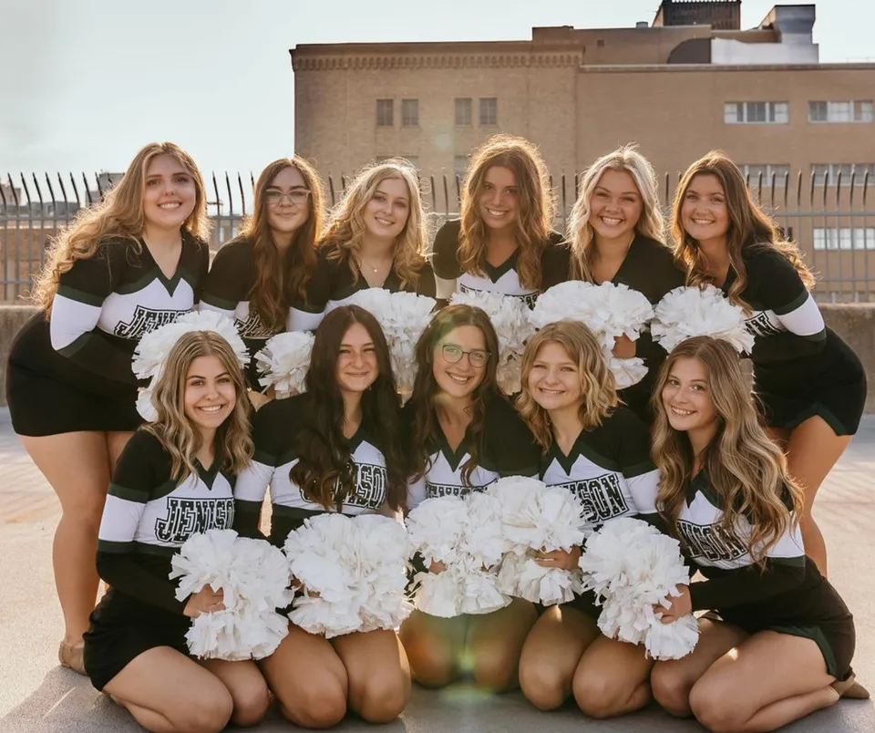 Un gruppo di giovani studentesse in uniformi da cheerleader con pon pon davanti a un edificio della Jenison High School.